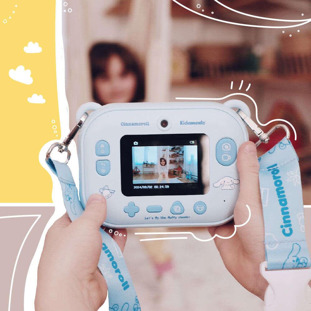 Close-up of a child holding the kiimento Model P Cinnamoroll camera, showing a preview screen with a girl posing for a photo. The lanyard strap with Cinnamoroll branding is visible, set against a cheerful background with illustrated accents.