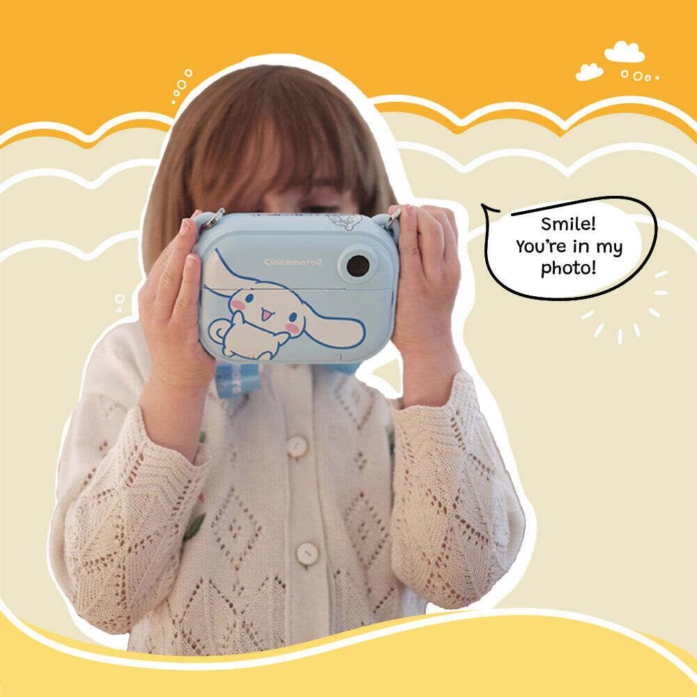 A young girl holds up the kiimento Model P Cinnamoroll kids’ digital camera as if taking a photo, with playful text reading “Smile! You’re in my photo!” on a cheerful yellow background with cartoon clouds.