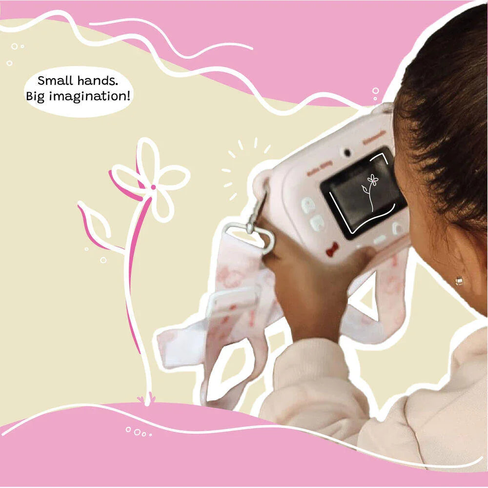 Close-up of a young girl using the kiimento Hello Kitty Model P camera to take a photo, viewed from behind. The camera screen displays a flower sketch, and the camera strap with Hello Kitty designs hangs around her hands. Caption reads “Small hands. Big imagination!” over a soft pink and beige illustrated background.